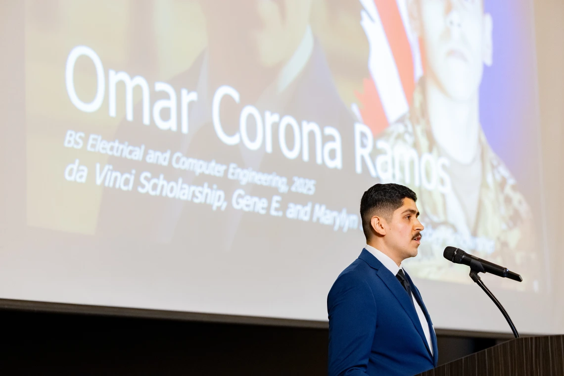 A young man with brown hair and a brown mustache reads a speech.