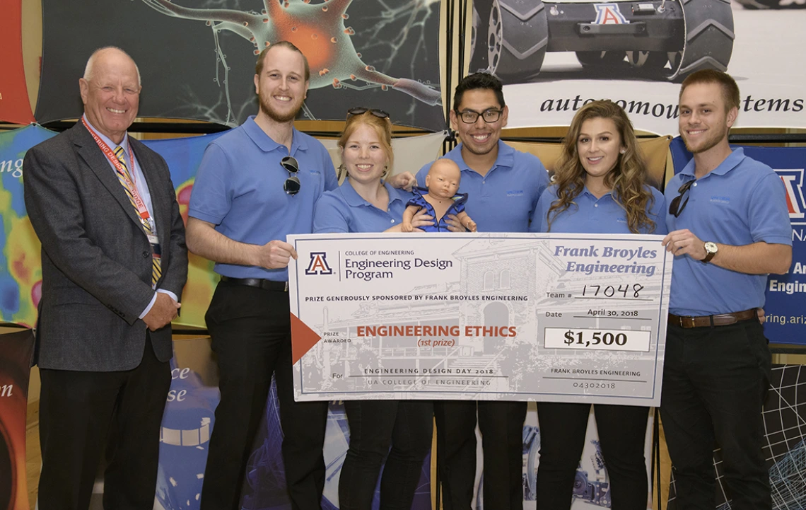 Team 17048 at Design Day 2018 A man in a suit standing next to five students wearing blue polo shirts and holding a giant check for $1,500