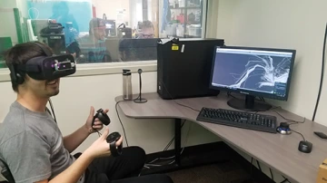 Team 17061 VR A student wearing VR goggles and handling two control pads looks around while a nearby monitor displays a computer-generated representation of a brain's white matter fiber tracts